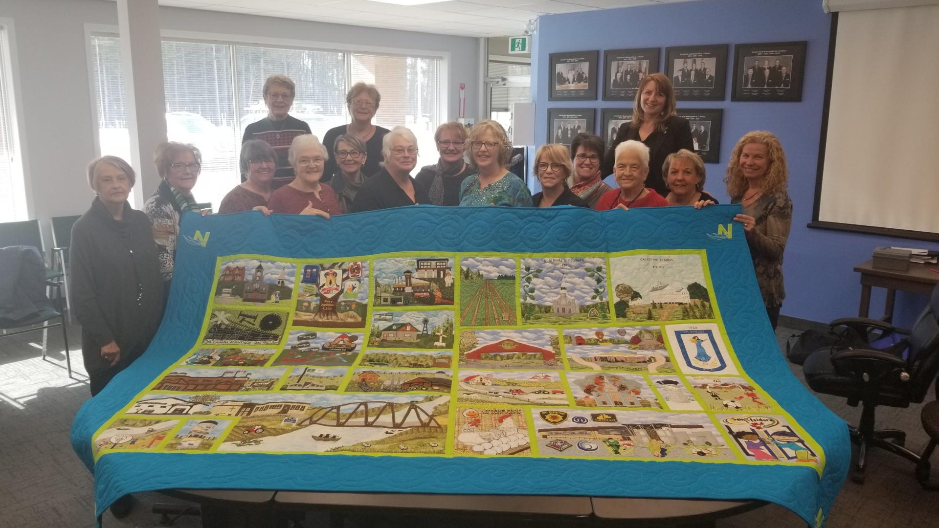 Group holding a large, colorful quilt in a room with framed photos on walls.