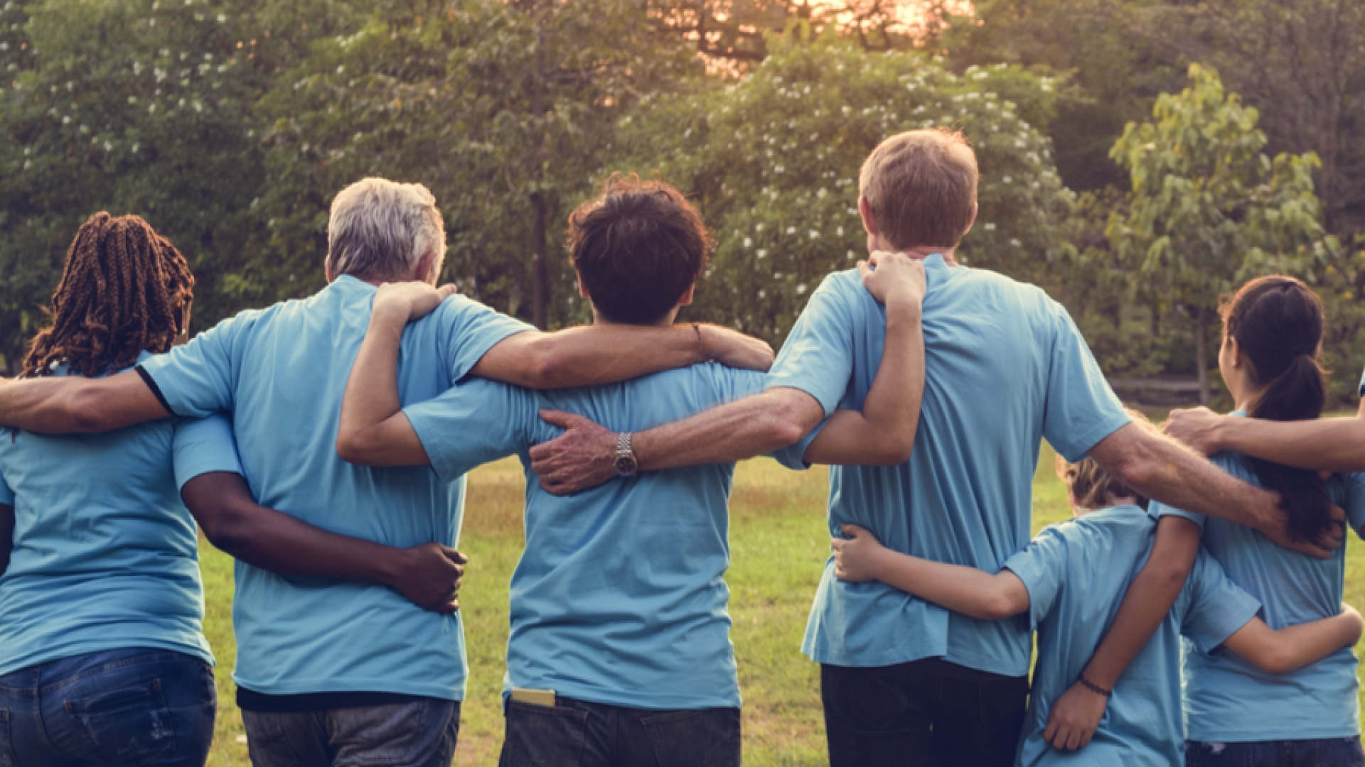 Seven people in blue shirts walk arm-in-arm in a park at sunset.