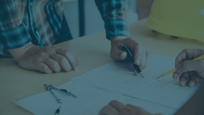 Two people review building plans and discuss a construction project at a table, with drafting tools and a hard hat nearby.