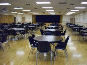 Empty dining hall with round tables and chairs, wooden floor, bright lighting.