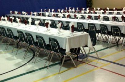 Rows of tables with white tablecloths, set with folded napkins, in a large hall.