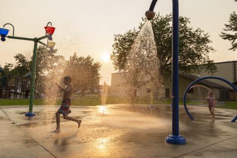 Children playing in a splash pad at sunset, water jets spraying around them.