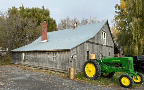 The historic Proulx Cheese Factory, a weathered wooden building surrounded by autumn trees, with a green tractor beside it.