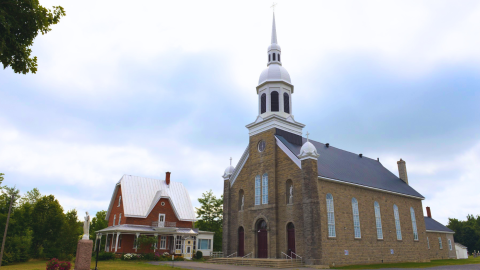 Stone church with tall white steeple beside a red brick house and statue, set against a cloudy sky and surrounded by greenery.
